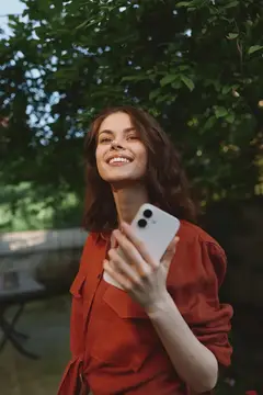 Улыбающаяся темноволосая девушка в красной блузе держит смартфон в саду