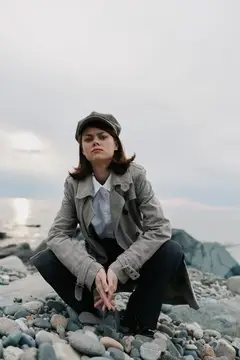 Молодая женщина в кепке и плаще сидит на камнях у моря