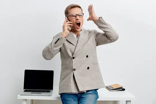 Мужчина в очках кричит в телефон у стола с ноутбуком