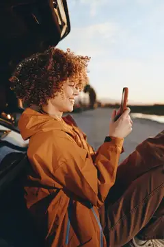 Женщина в стиле лайфстайл со смартфоном в машине, портрет с улыбкой на закате, вьющиеся волосы ловят золотой свет