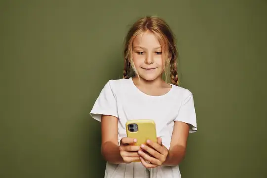Светловолосая девочка с косичками смотрит в жёлтый смартфон на зелёном фоне