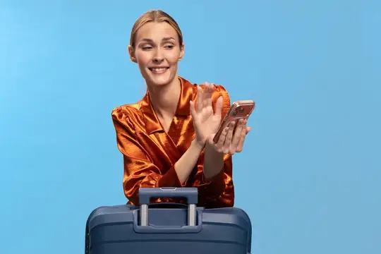 Счастливая путешественница в яркой блузке машет рукой со смартфоном в руке, стоя за чемоданом на голубом фоне