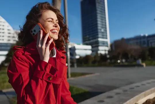 Улыбающаяся кудрявая девушка в красном плаще говорит по телефону на городской улице