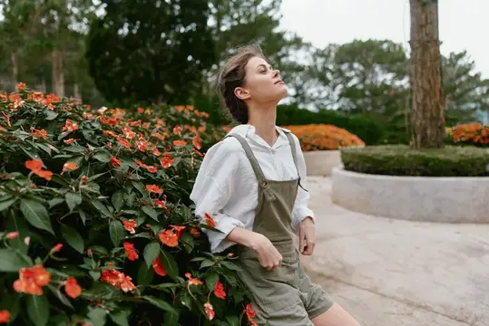 Веселая молодая женщина в повседневной одежде стоит среди красочных цветов, наслаждаясь природой с безмятежным выражением лица