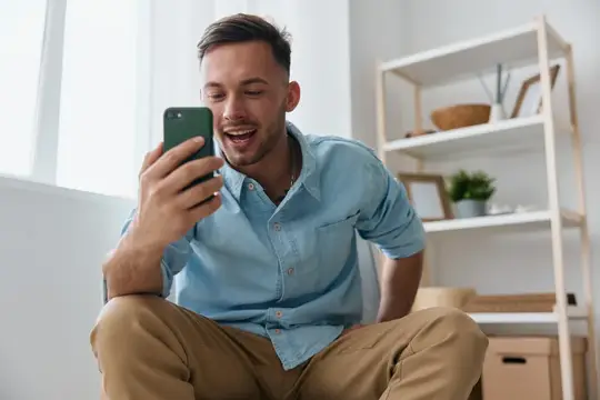 Улыбающийся мужчина в голубой рубашке смотрит в смартфон дома