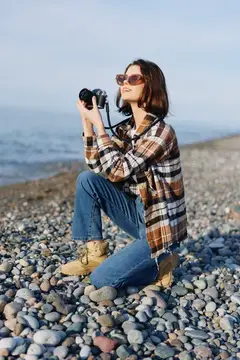 Сцена уличной фотографии: женщина с камерой на галечном берегу, одетая в клетчатую рубашку и джинсы, присела и снимает морской пейзаж с сосредоточенностью и стилем