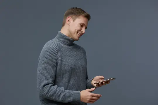 Улыбающийся мужчина в сером свитере смотрит в смартфон на сером фоне
