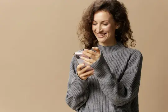 Улыбающаяся кудрявая женщина в сером свитере смотрит в телефон