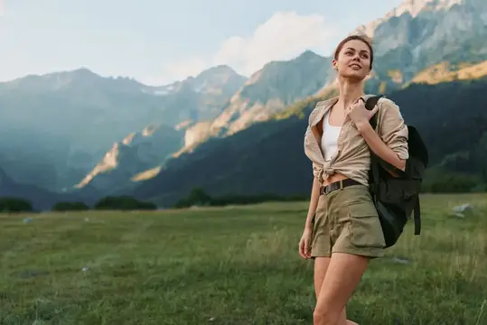 Улыбающаяся женщина в шортах и завязанной узлом рубашке любуется горным пейзажем, держась за лямку рюкзака, что иллюстрирует концепции пешего туризма, путешествий и активного образа жизни