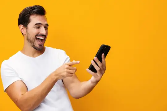 Смеющийся темноволосый мужчина показывает пальцем на смартфон на жёлтом фоне