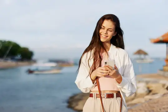 Улыбающаяся темноволосая девушка со смартфоном на берегу моря
