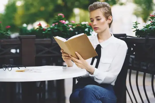 Девушка с короткой стрижкой читает книгу за столиком летнего кафе