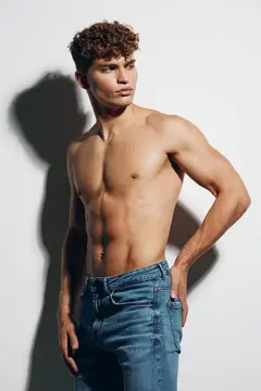 Кудрявый парень с обнажённым торсом позирует в джинсах на светлом фоне