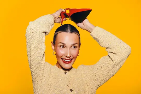 Улыбающаяся женщина держит красную туфлю над головой на жёлтом фоне