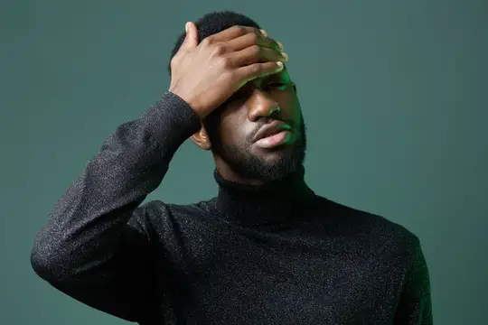 Темнокожий мужчина в черной водолазке закрыл лицо рукой на зеленом фоне