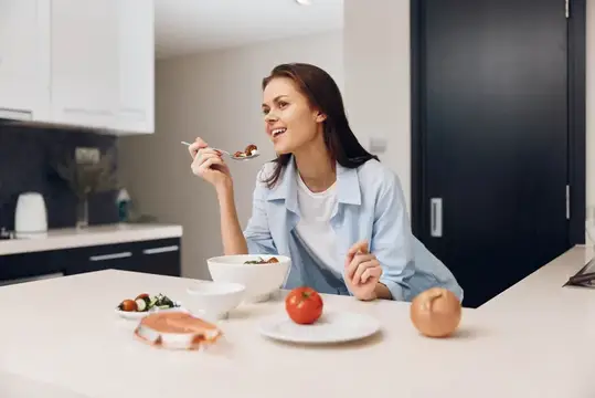 Улыбающаяся женщина ест салат за столом на светлой кухне