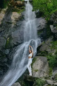 Молодая женщина в белом стоит на камне у подножия водопада