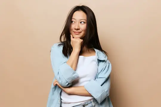 Милый, улыбающийся, задумчивый, загорелый, восхитительный молодой азиатка опирается на руку, сжав кулак, и позирует изолированно на бежевом пастельном фоне