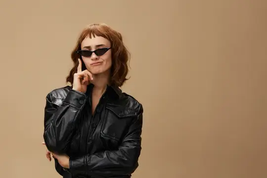 Рыжеволосая девушка в солнечных очках и кожаной куртке задумчиво смотрит в сторону