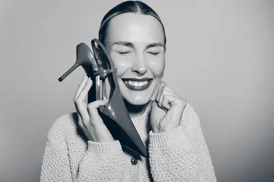 Улыбающаяся женщина прижимает туфлю к уху как телефон