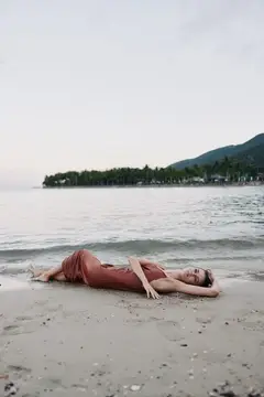Мирная женщина отдыхает на песчаном берегу у спокойной воды с пышными зелеными холмами на заднем плане под мягким пастельным небом на закате