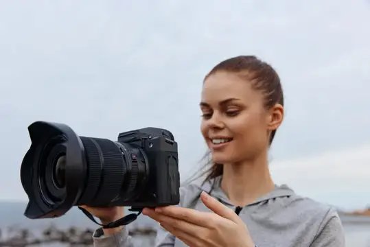 Веселая молодая женщина в повседневной серой одежде держит фотоаппарат на пляже в окружении спокойного океанского пейзажа