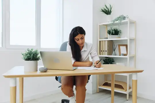 Женщина работает и учится, записывая текст на бумаге ручкой и проверяя ошибки, занимаясь учебой и преподаванием в колледже, выполняя деловую работу за столом с ноутбуком