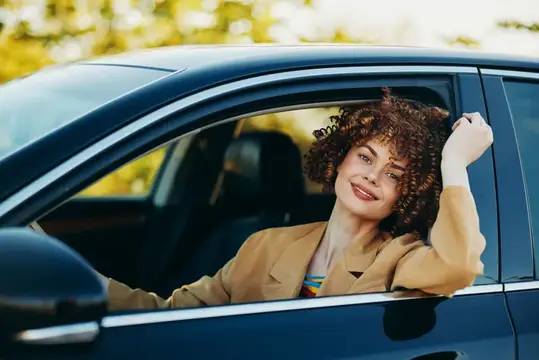 Кудрявая девушка в бежевом пиджаке выглядывает из окна автомобиля