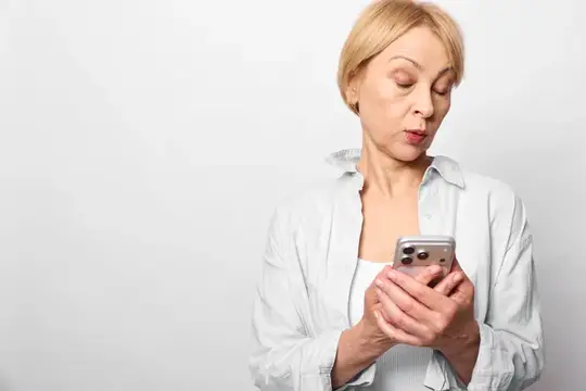Блондинка в белой рубашке с интересом смотрит в смартфон на светлом фоне