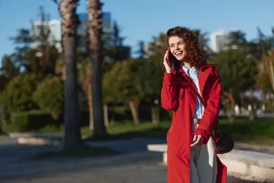 Улыбающаяся кудрявая девушка в красном плаще говорит по телефону на улице