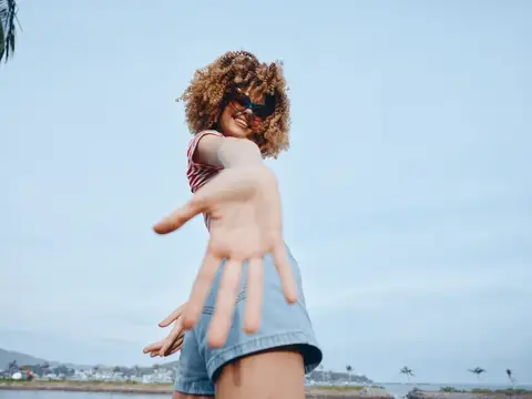 Улыбающаяся кудрявая девушка в очках тянет руку к камере на фоне моря
