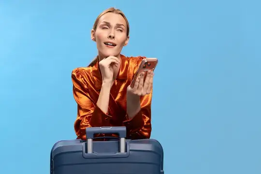 Девушка в оранжевой блузе смотрит в телефон, опираясь на чемодан