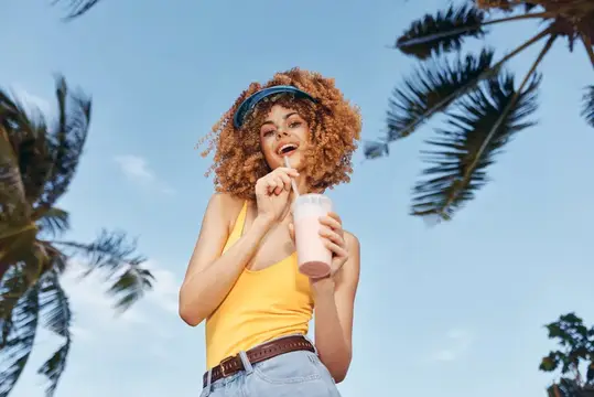 Кудрявая девушка в жёлтом топе пьёт коктейль на фоне пальм