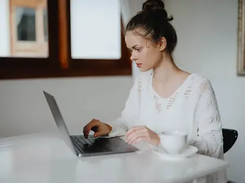 Молодая женщина в белом свитере работает за ноутбуком с чашкой кофе