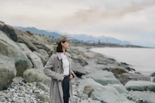 Молодая женщина в плаще и кепке стоит на каменистом берегу