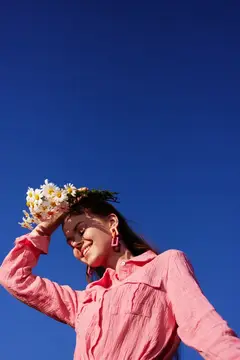 Веселая молодая женщина в розовой рубашке и цветочном венке выражает радость на фоне ярко-голубого неба, воплощая дух весны и красоту природы