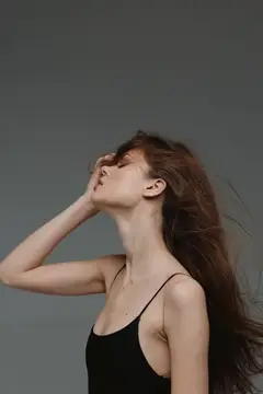 Профильный портрет молодой женщины с длинными развевающимися волосами и в черном топе, которая грациозно позирует с закрытыми глазами на простом сером фоне