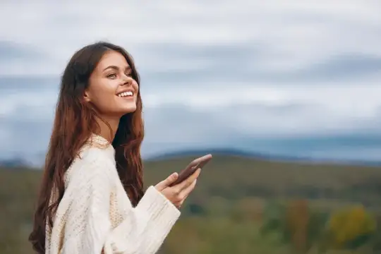 Улыбающаяся темноволосая девушка со смартфоном на фоне холмов