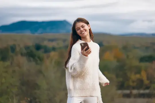 Улыбающаяся девушка в белом свитере смотрит в телефон на фоне гор