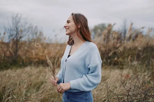 Улыбающаяся девушка в голубом кардигане стоит в осеннем поле