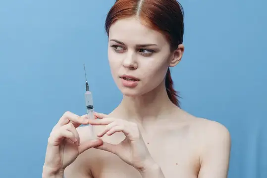 Женщина делает укол, её голые плечи и испуганное лицо на синем фоне