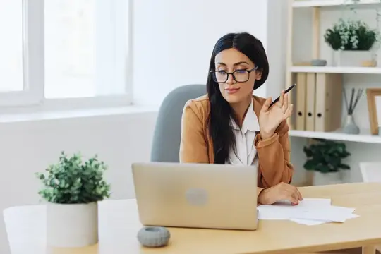 Бизнес-леди в бежевом костюме и белой рубашке в очках работает в офисе за своим столом с ноутбуком, проводит онлайн-видеочат, преподаватель занимается со студентами