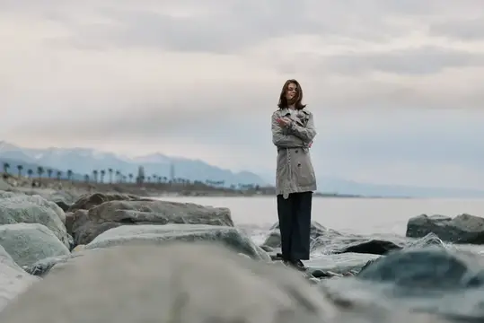 Девушка в плаще стоит на камнях у моря в пасмурный день