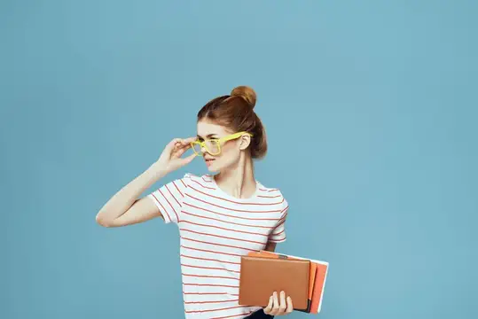 Студентка с книгами в руках на голубом фоне, в жёлтых очках, с модельной причёской, кадрированный вид