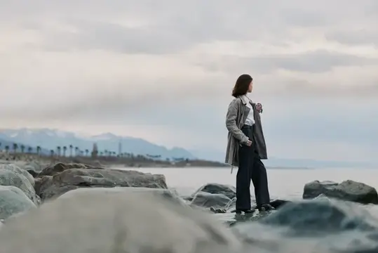 Молодая женщина в плаще стоит на камнях у моря в пасмурный день