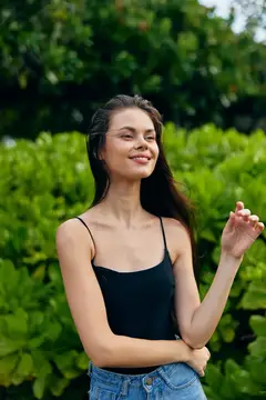 Улыбающаяся темноволосая девушка в чёрном топе на фоне зелёных кустов