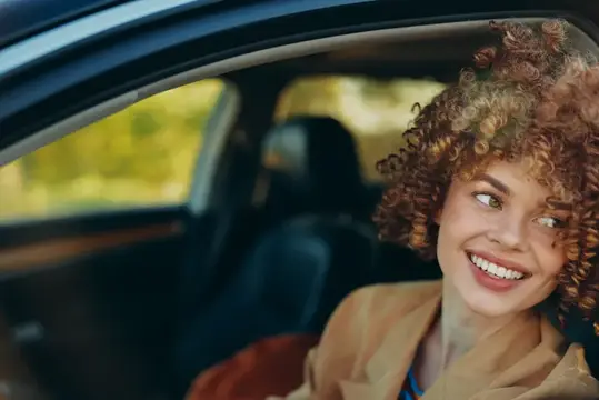 Рыжеволосая девушка с кудрями улыбается, выглядывая из окна автомобиля