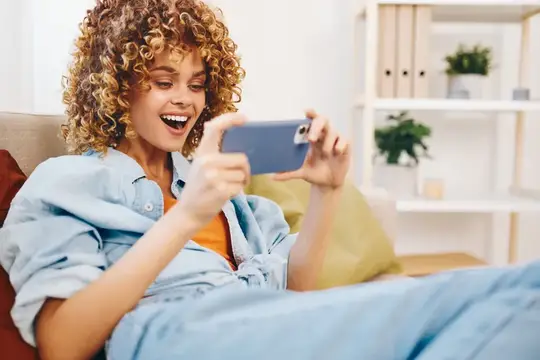 Кудрявая девушка смеётся, глядя в смартфон, лёжа на диване