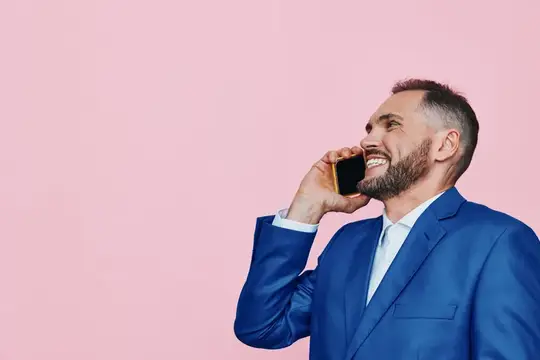 Улыбающийся бородатый мужчина в синем костюме говорит по телефону на розовом фоне
