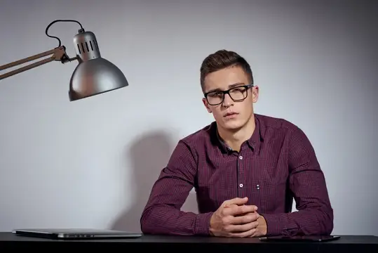 Деловой мужчина в очках и рубашке сидит за столом в темной комнате с лампой, обрезанный вид